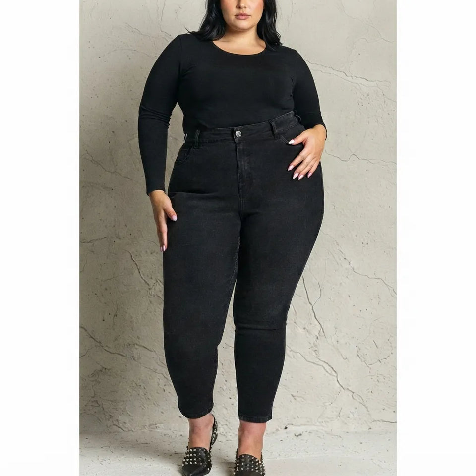 A plus-size model wearing black high-waisted skinny jeans with a matching black belt, styled with a black long-sleeve top and black studded heels, standing against a textured light gray wall.