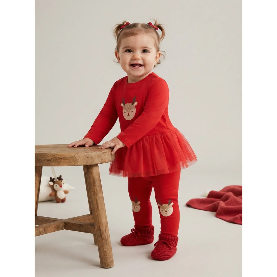 A toddler girl wearing a festive red holiday outfit with a long-sleeve top, tutu skirt, matching leggings, and knit booties, standing next to a wooden stool against a light gray background.