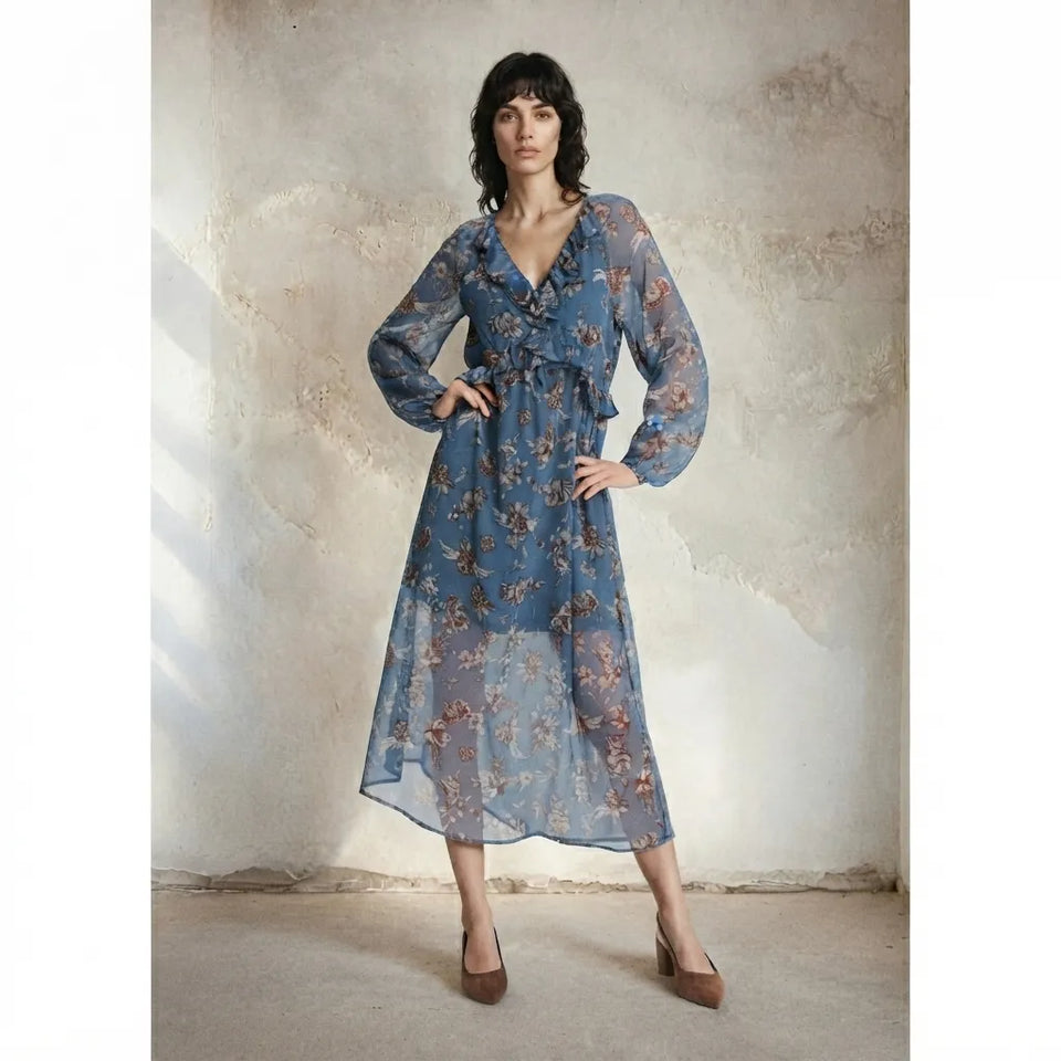 A woman models a long-sleeved, blue floral midi dress with a tie waist, standing against a textured plaster wall.