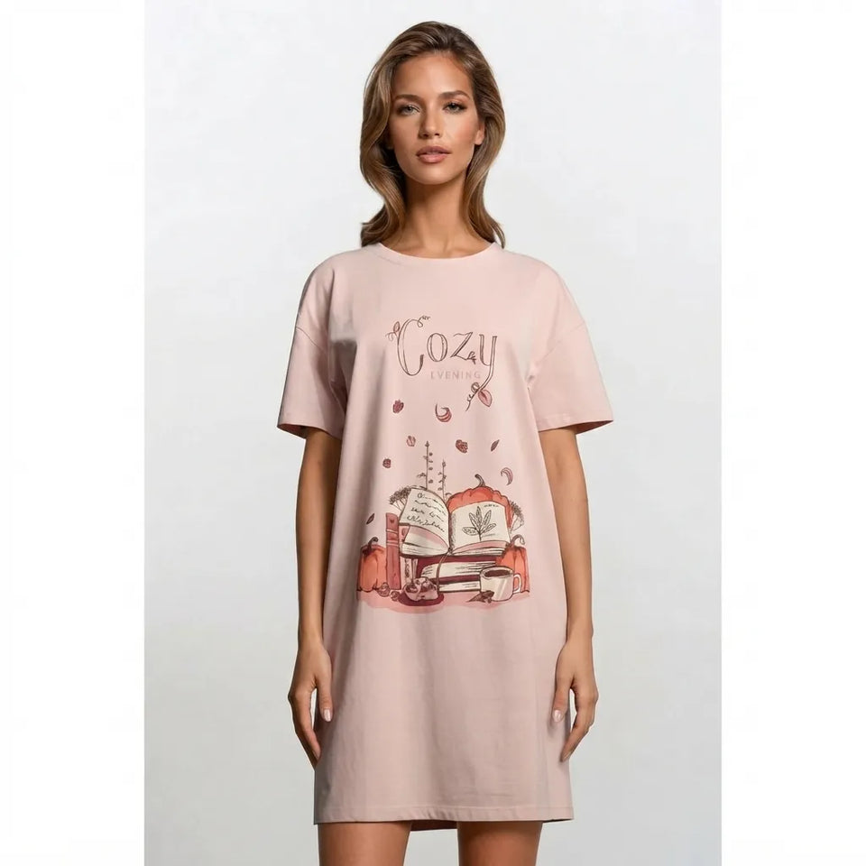 A woman modeling a light pink short-sleeve t-shirt dress with a vintage-style graphic on the front featuring the text 'Cozi' and an illustration of a book and coffee cups.
