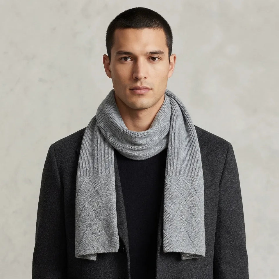 A man wearing a light gray ribbed knit scarf wrapped around his neck, styled with a dark gray blazer and black shirt against a textured light gray background.