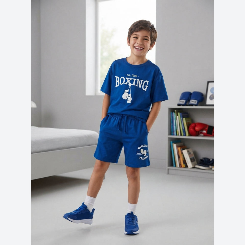 A smiling boy stands in a bright room wearing a royal blue boxing-themed t-shirt and matching shorts with white graphics, white socks, and blue sneakers.