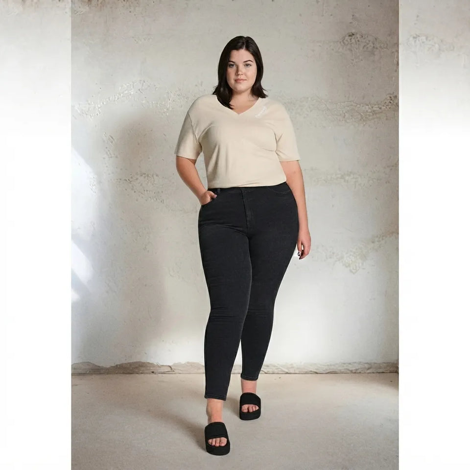 A woman modeling a beige short-sleeve V-neck top and black skinny jeans against a textured concrete wall.