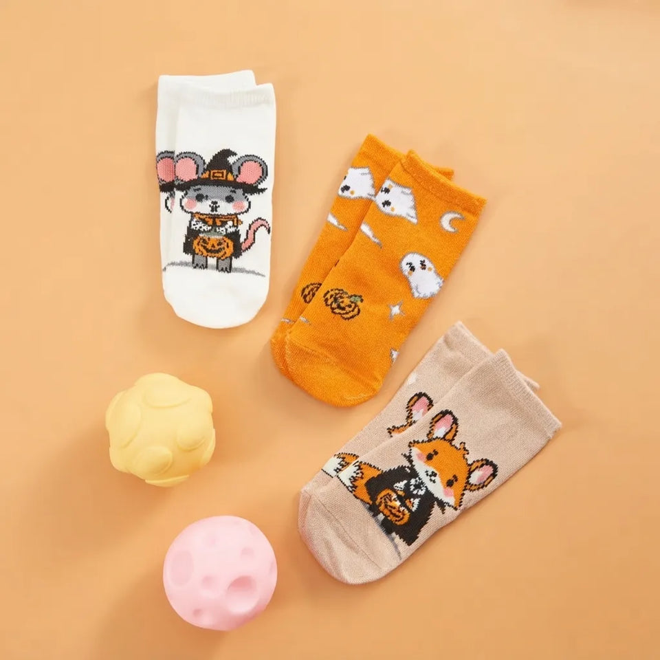 Three pairs of ankle socks with cute cartoon mouse Halloween designs, displayed on a warm peach background alongside two small decorative bath bombs.