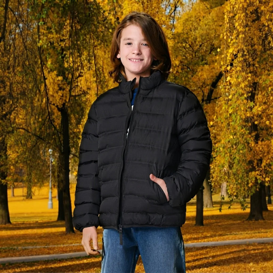 A young boy wearing a black puffer jacket and blue jeans, standing in a park with autumn trees in the background.