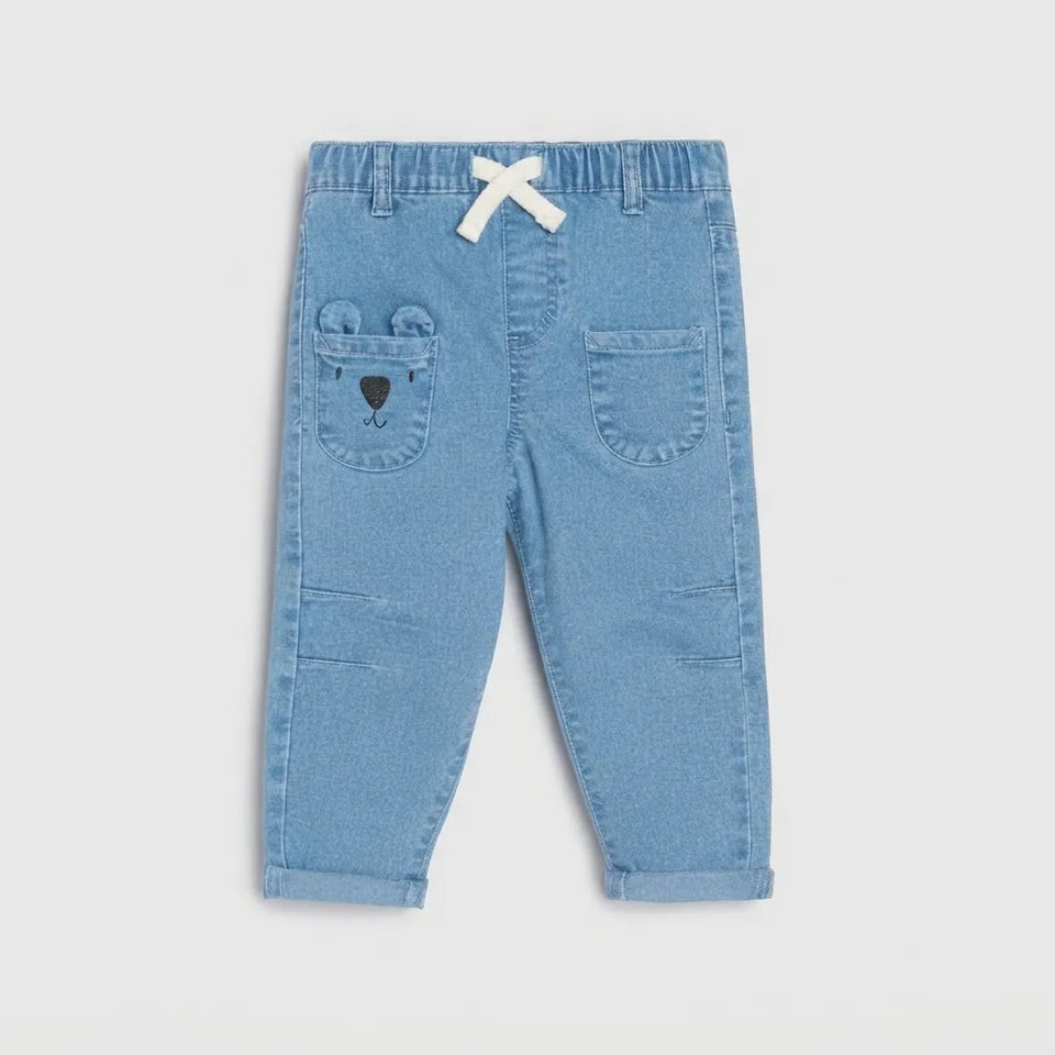 Light blue denim toddler pants with a white bow at the elastic waist, a bear face detail on the back pocket, and side utility pockets, shown flat on a light gray background.