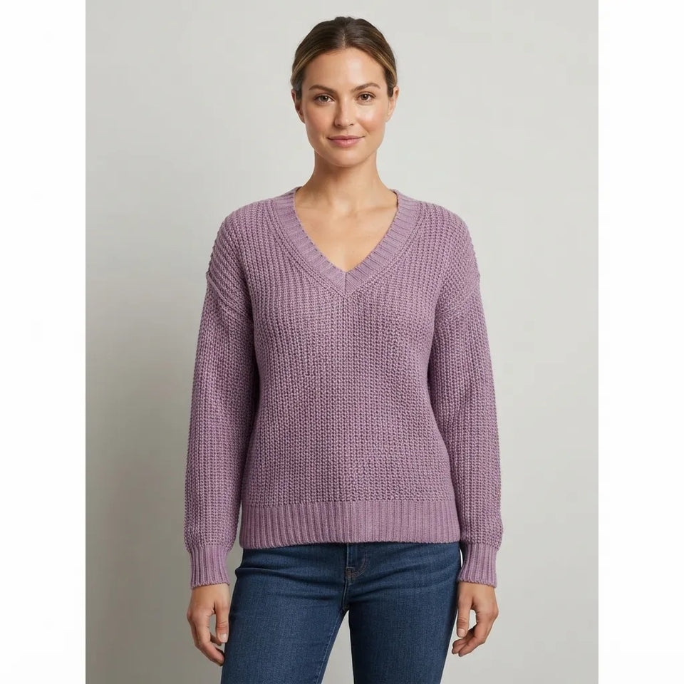 A woman wearing a textured, lavender-purple V-neck knit sweater and dark blue jeans, standing against a neutral light gray background.