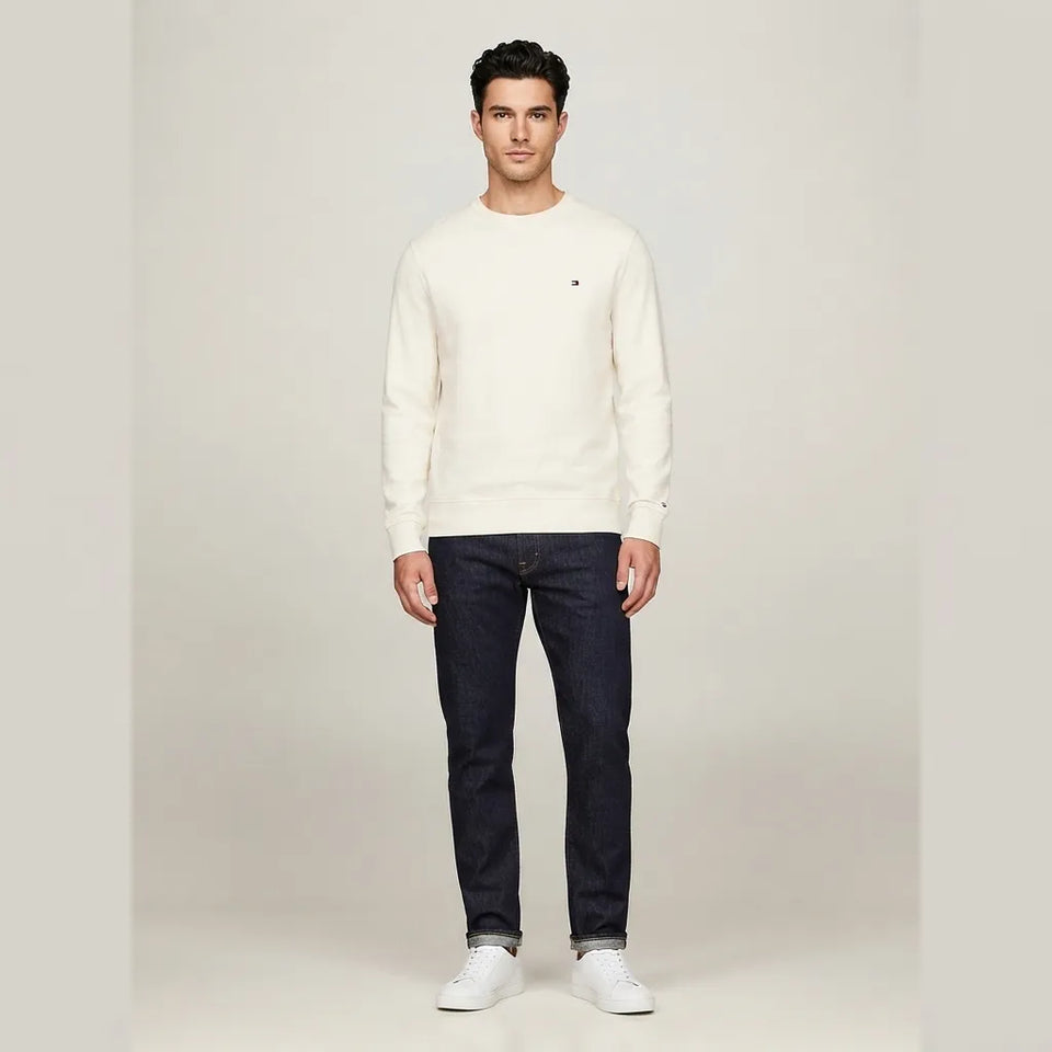 A man modeling a cream crewneck sweater with a small Tommy Hilfiger flag logo on the left chest, paired with dark blue jeans and white sneakers, against a light gray studio background.