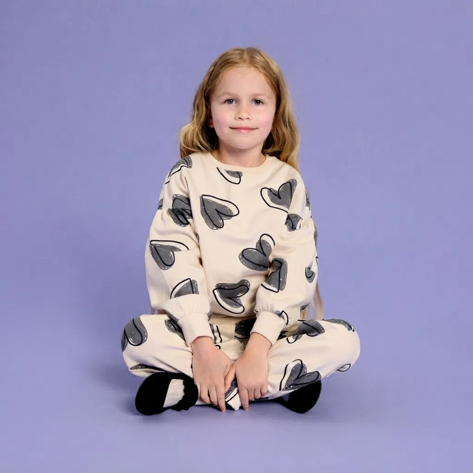 A young girl with blonde hair sits cross-legged on a solid lavender studio background, wearing a cream-colored two-piece pajama set with a dark gray heart pattern.