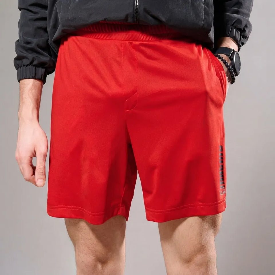 Red athletic shorts worn by a model, shown from the waist down against a neutral gray studio background. The model is also wearing a black zip-up jacket and a black watch.