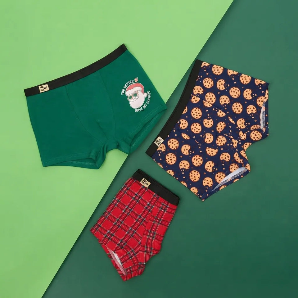 Three pairs of kids' boxer briefs laid flat on a split green background. One pair is dark green with a black waistband and a Santa graphic; one pair is navy with a cookie print; one pair is red plaid with a black waistband.