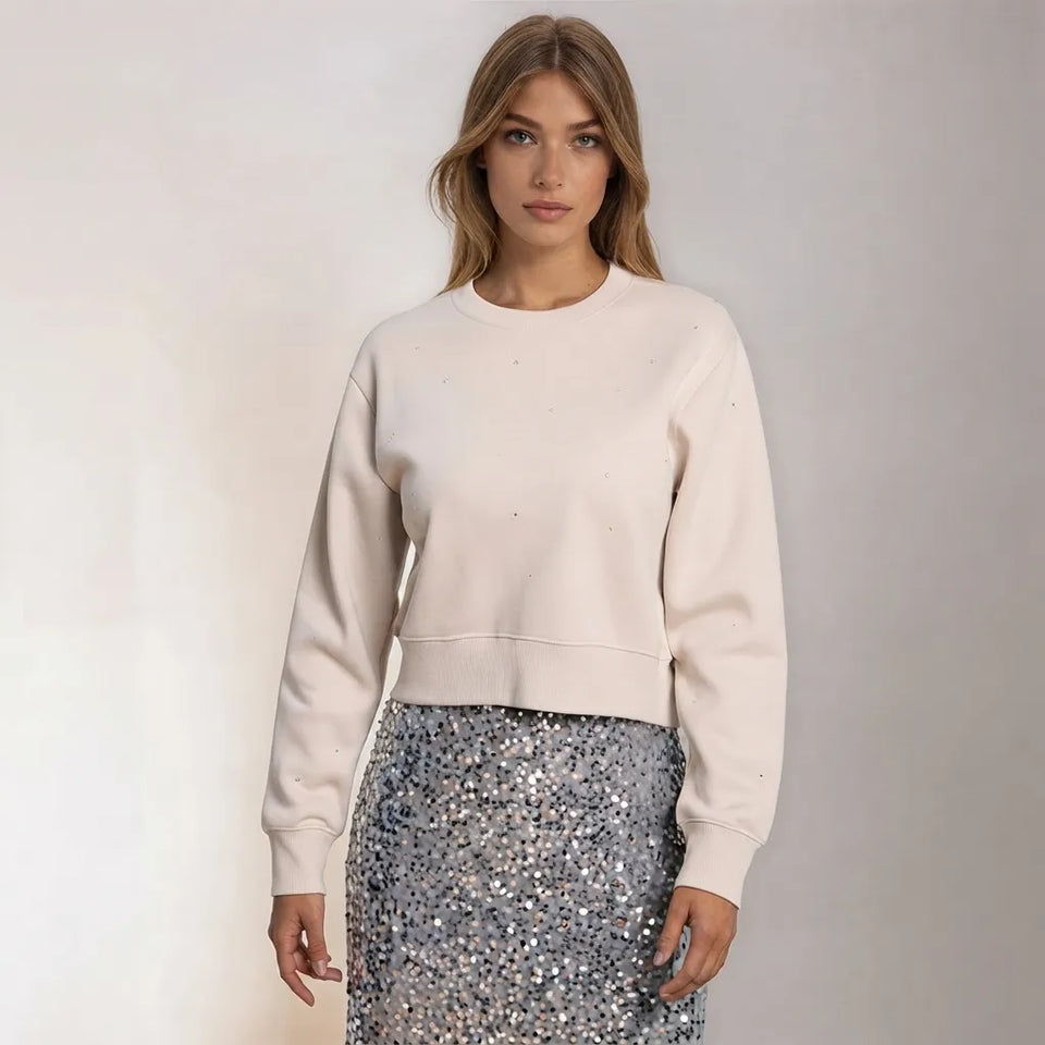 A woman modeling a light cream crewneck sweatshirt paired with a silver sequined skirt, set against a plain light gray studio background.