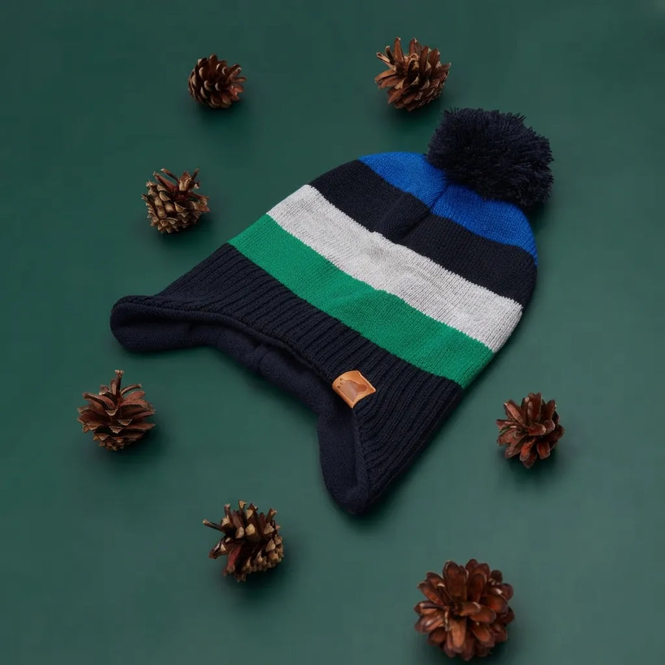 A striped knit beanie with a pom-pom, laid flat on a dark green background and surrounded by scattered pinecones. The hat has horizontal stripes in navy, blue, white, and green, with a small tan leather patch near the ear flap.