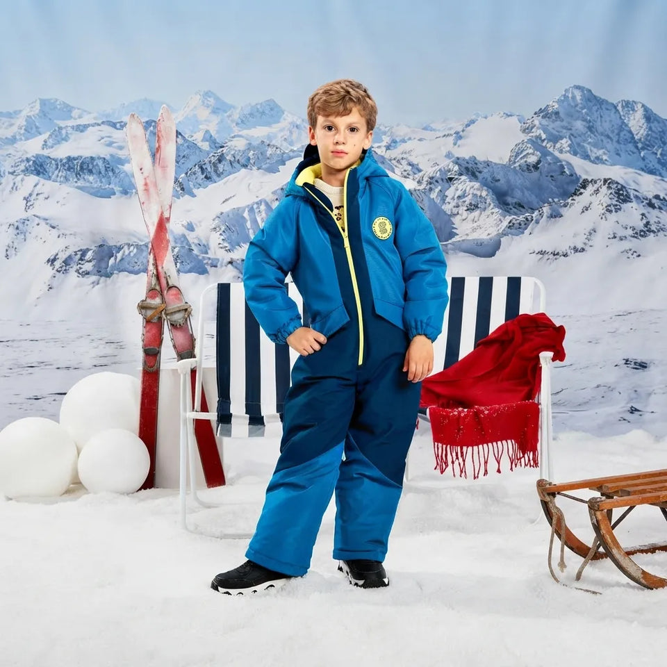 A child wearing a bright blue ski suit with yellow zipper accents stands in a snowy outdoor-style set with mountains in the background, skis, a striped chair, a red fringed blanket, and a sled.
