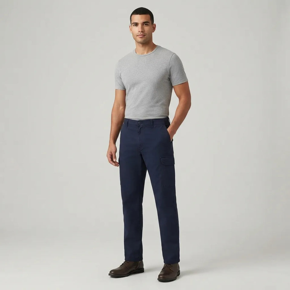 A man modeling navy blue trousers with a light gray t-shirt and brown leather shoes, standing against a light gray studio background.