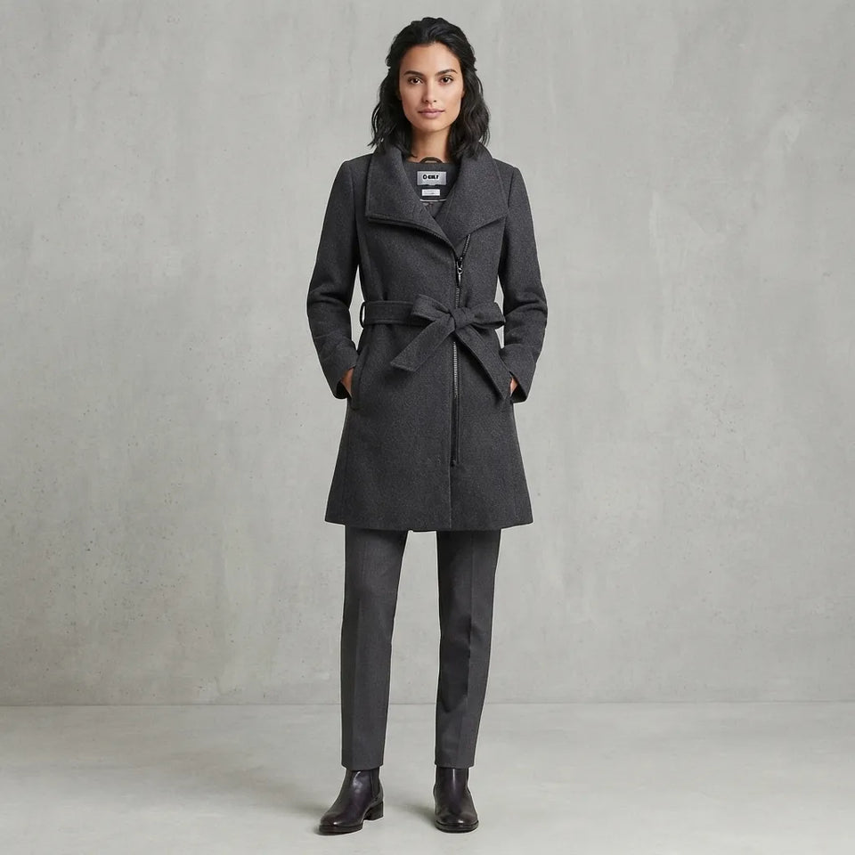 A woman stands against a textured gray studio backdrop wearing a dark charcoal gray belted coat over a matching gray suit with dark brown ankle boots.