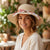 A woman wearing a wide-brimmed straw sun hat with a red, white, and blue patterned silk ribbon band tied around the crown, set against a warm, softly blurred outdoor patio background.