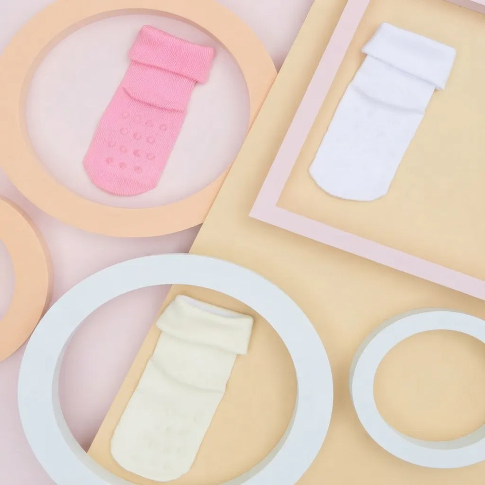 Three soft fabric baby socks in pink, white, and cream are arranged on pastel geometric paper circles and frames.