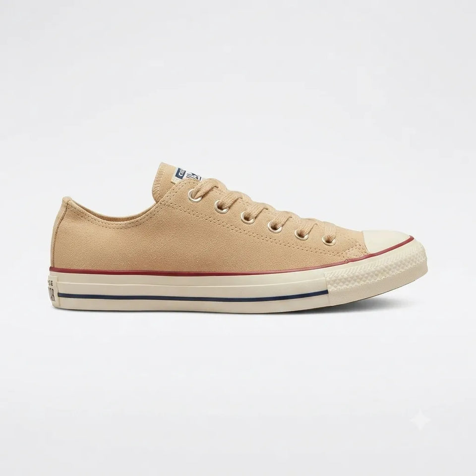 A classic low-top canvas sneaker in a light tan/beige color, featuring a cream rubber toe cap, a thin red stripe above the sole, and a navy blue stripe below it, set against a plain white background.