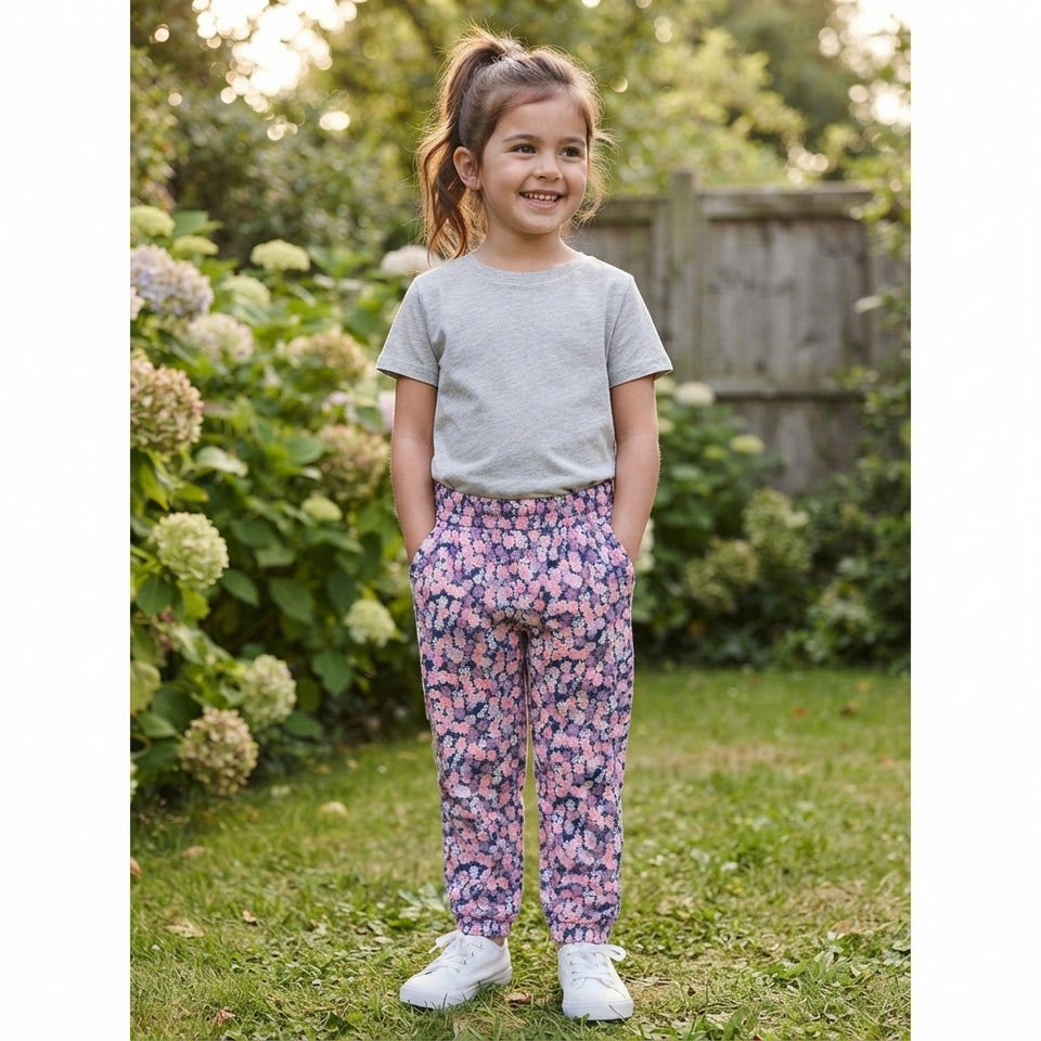 A young girl stands outdoors in a garden wearing a light gray short-sleeve t-shirt and pink and purple floral print pants, paired with white sneakers.