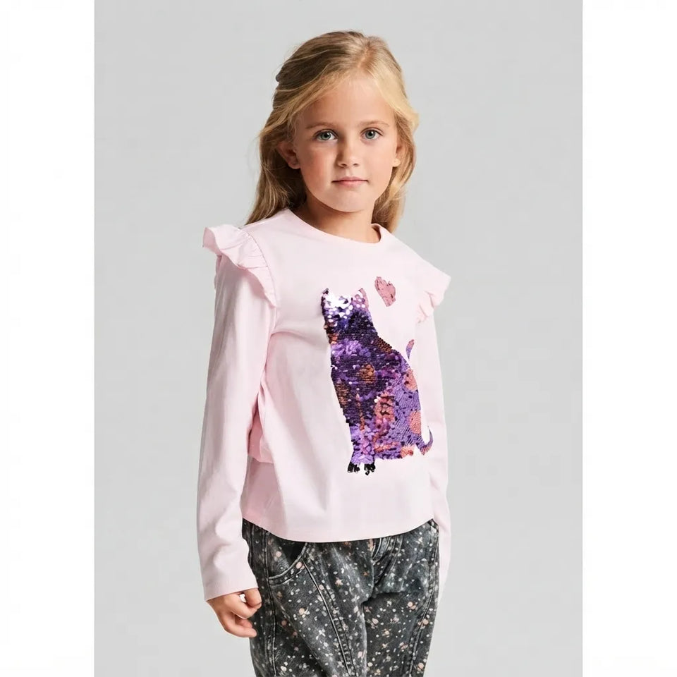 A young girl with blonde hair wearing a light pink long-sleeve top with ruffled shoulders and a purple sequined cat graphic, paired with dark patterned pants, against a light gray background.