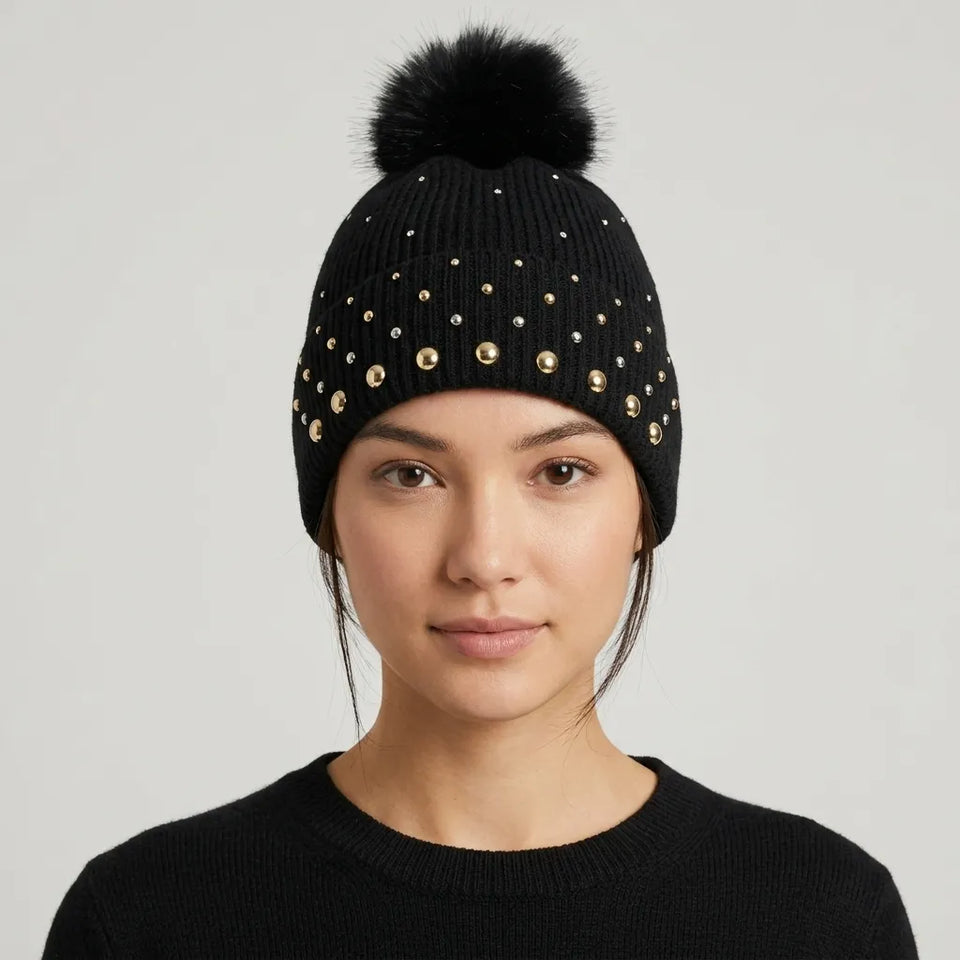 A woman wearing a black knit beanie with a large black pom-pom and gold-tone studs, shown from the chest up against a light gray studio background.