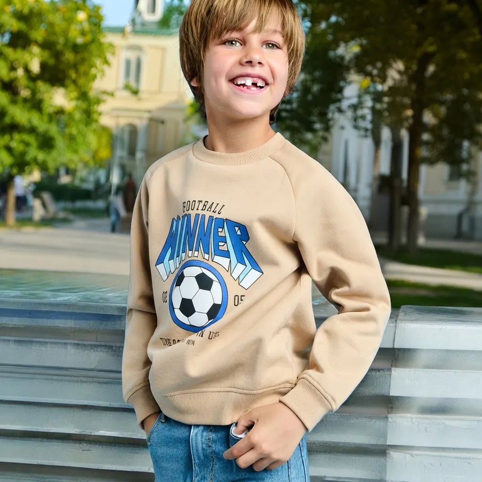 A young boy smiling while wearing a beige crewneck sweatshirt with a blue and white graphic that reads 'FOOTBALL WINNER' around a soccer ball, paired with blue jeans, set against an outdoor background of trees and buildings.