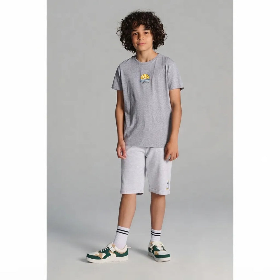 A child model wearing a heather gray short-sleeve t-shirt with a small yellow graphic on the chest, matching light gray shorts, white socks, and green and white sneakers, standing against a neutral gray studio background.
