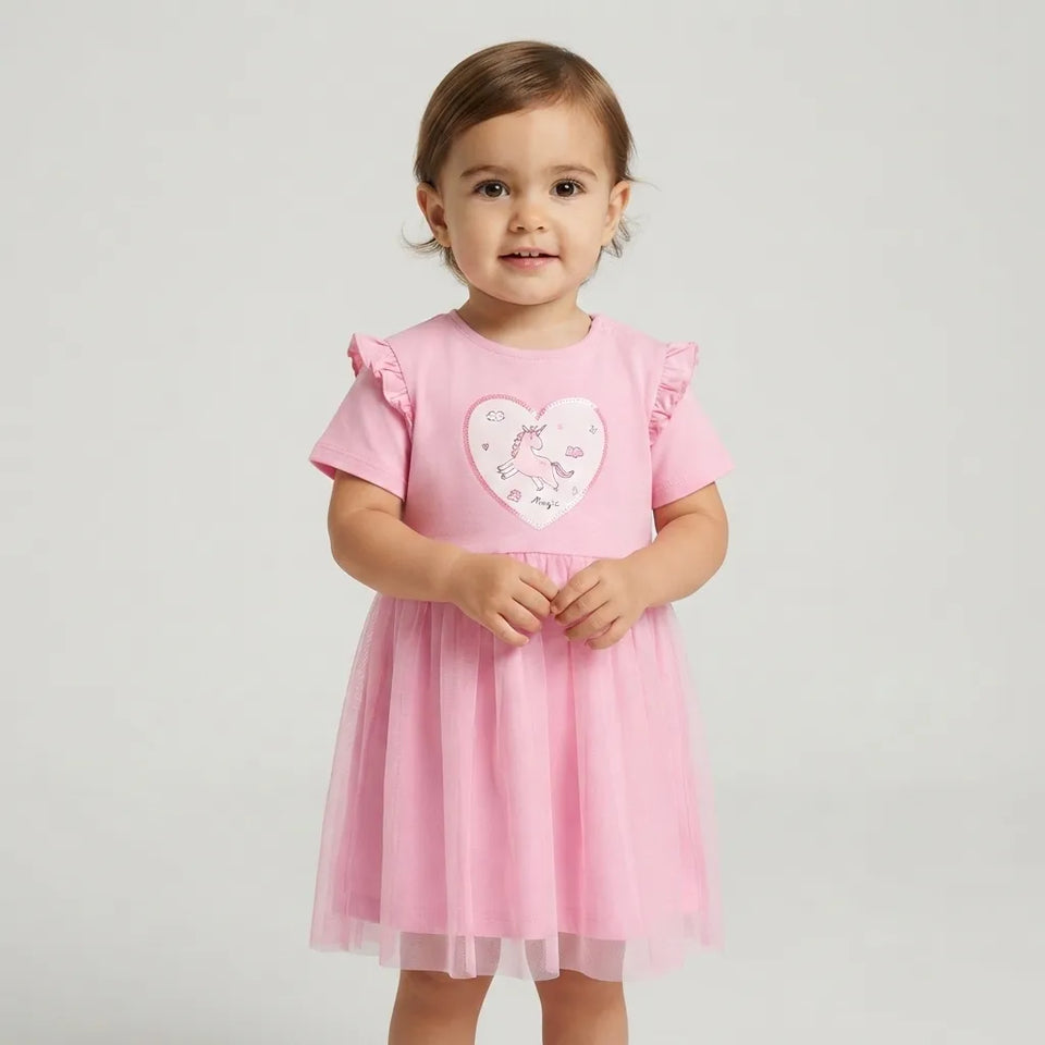 A toddler girl wearing a pink short-sleeve dress with ruffled shoulders and a tulle skirt, featuring a heart-shaped applique on the chest, photographed against a light gray studio background.