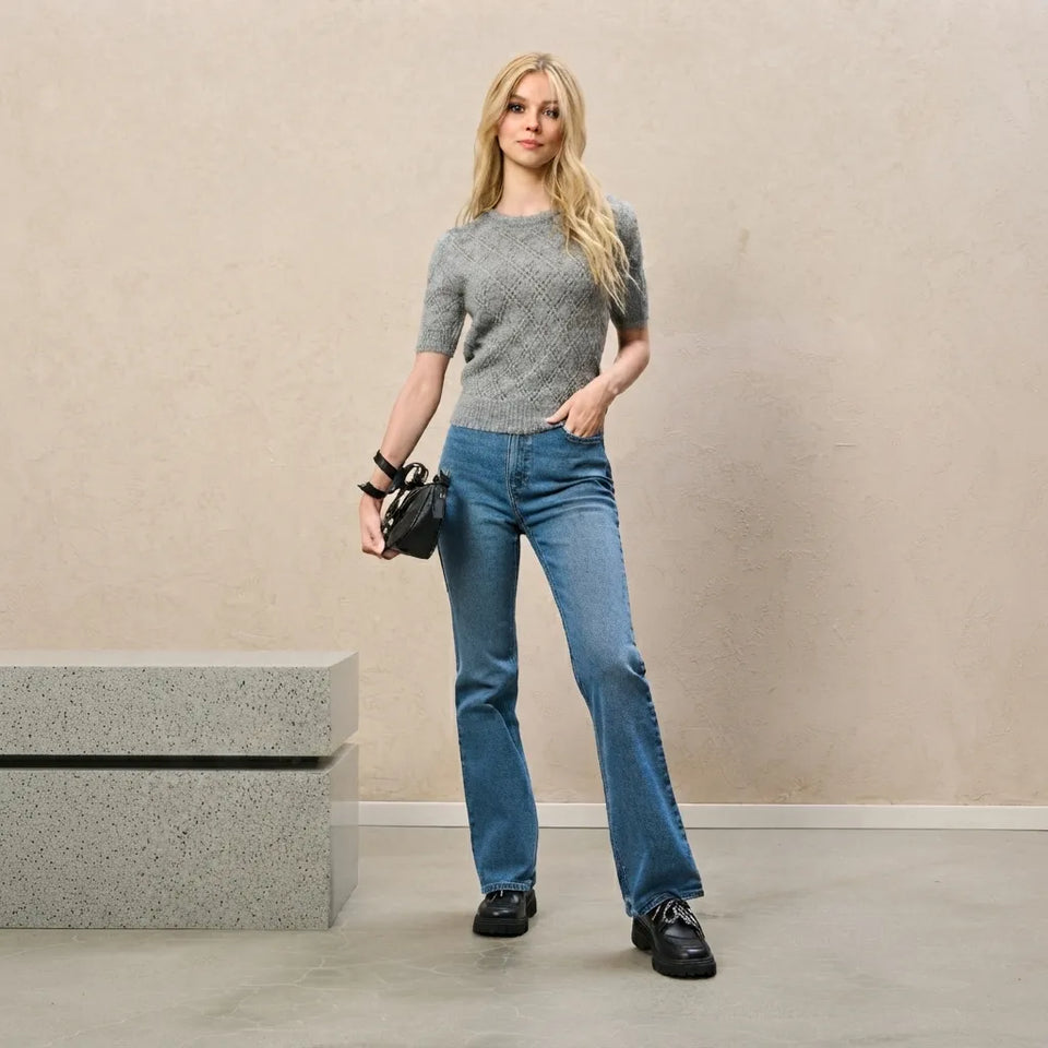 A woman stands in a studio wearing a gray short-sleeve knit top, blue flare jeans, and black shoes, holding a small black handbag. The background is a warm beige textured wall with a concrete bench on the left.
