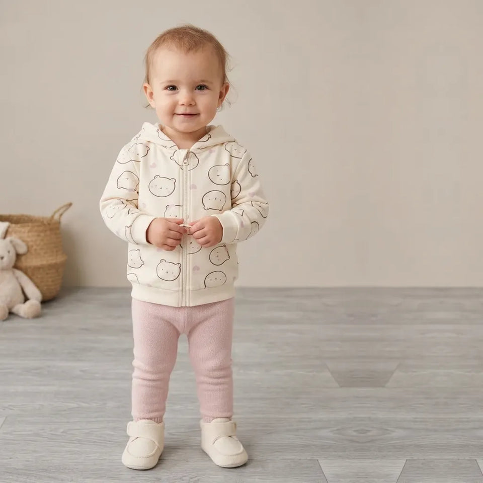 A baby standing in a room wearing a cream zip-up hoodie with a black cat pattern, pink pants, and cream shoes.