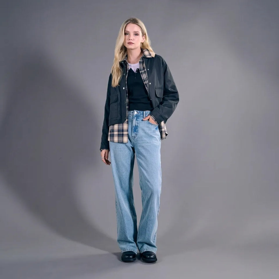 A blonde model stands against a gray studio background wearing a black overshirt over a dark top, a plaid shirt tucked into light-wash wide-leg jeans, and black shoes.