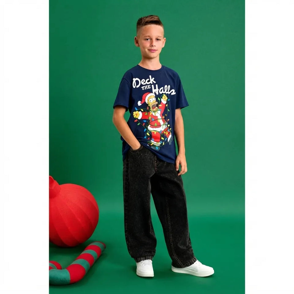 A young boy modeling a navy blue graphic t-shirt that reads 'Deck the Halls' with a festive Santa Claus illustration, paired with dark jeans and white sneakers, standing against a solid green studio backdrop.