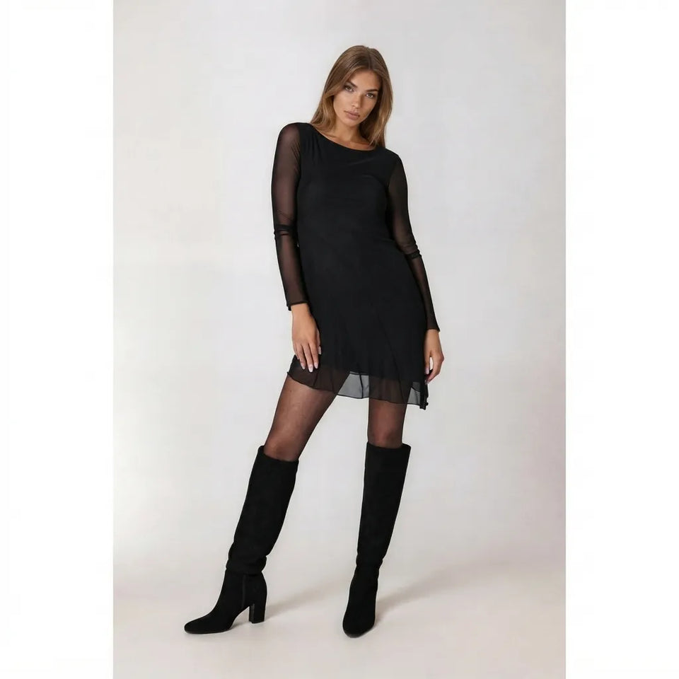 A woman modeling a black long-sleeve mini dress with sheer mesh sleeves and a sheer hem overlay, paired with black tights and black knee-high boots, against a plain light gray studio background.