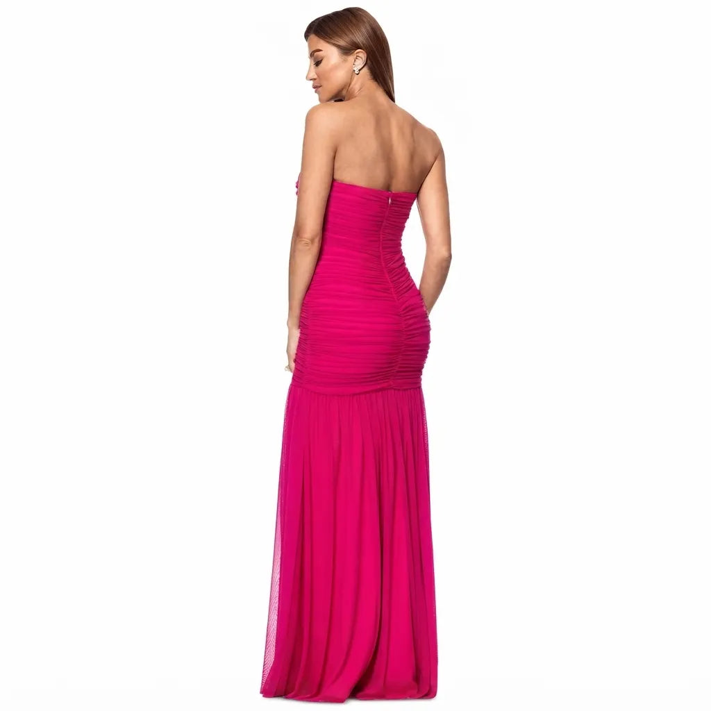 A woman modeling a strapless magenta pink maxi gown from the back, showing a fitted ruched bodice and a flowing skirt, set against a plain white background.