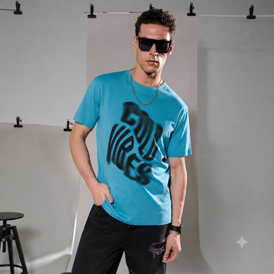 A man modeling a bright turquoise blue t-shirt with a black spray-paint style graphic reading 'COOL VIBES' on the front, paired with black pants and sunglasses in a studio setting.