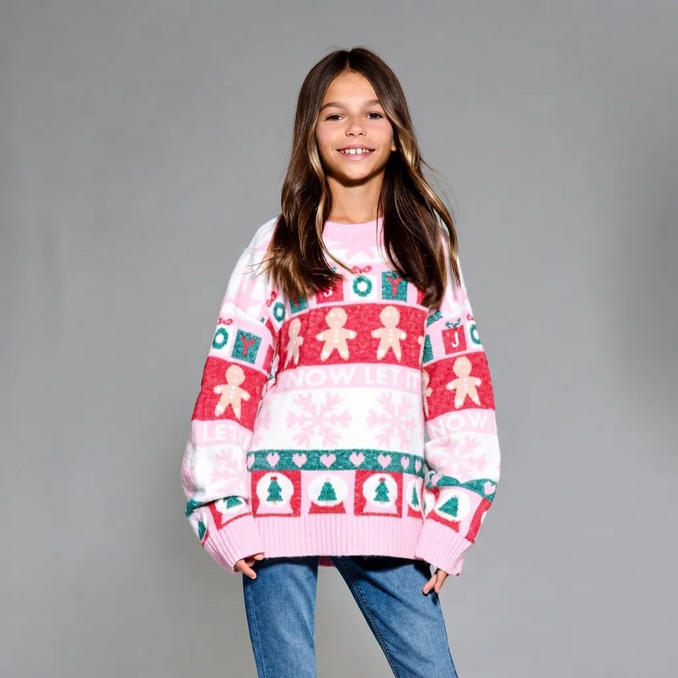 A young girl smiling while wearing a pink, red, and green Christmas knit sweater with gingerbread men and snowflake patterns, paired with blue jeans, against a solid gray studio background.