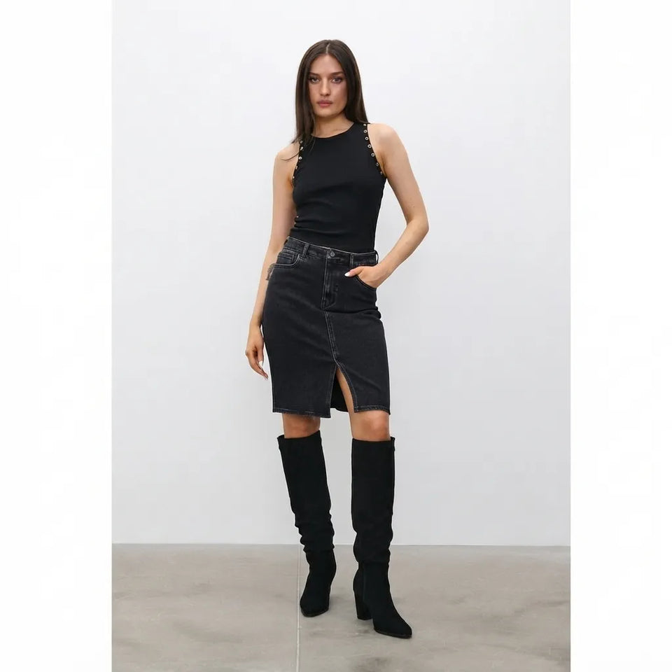 A woman modeling a black sleeveless top with decorative shoulder straps, a black denim midi skirt with a front slit, and black over-the-knee boots, standing against a light gray studio background.
