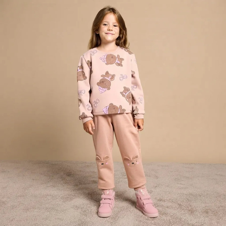 A young girl stands against a plain beige studio backdrop wearing a matching two-piece pajama set. The set is light pink with a brown bunny print on the long-sleeve top and matching pink pants featuring bunny ears and bunny faces on the knees.