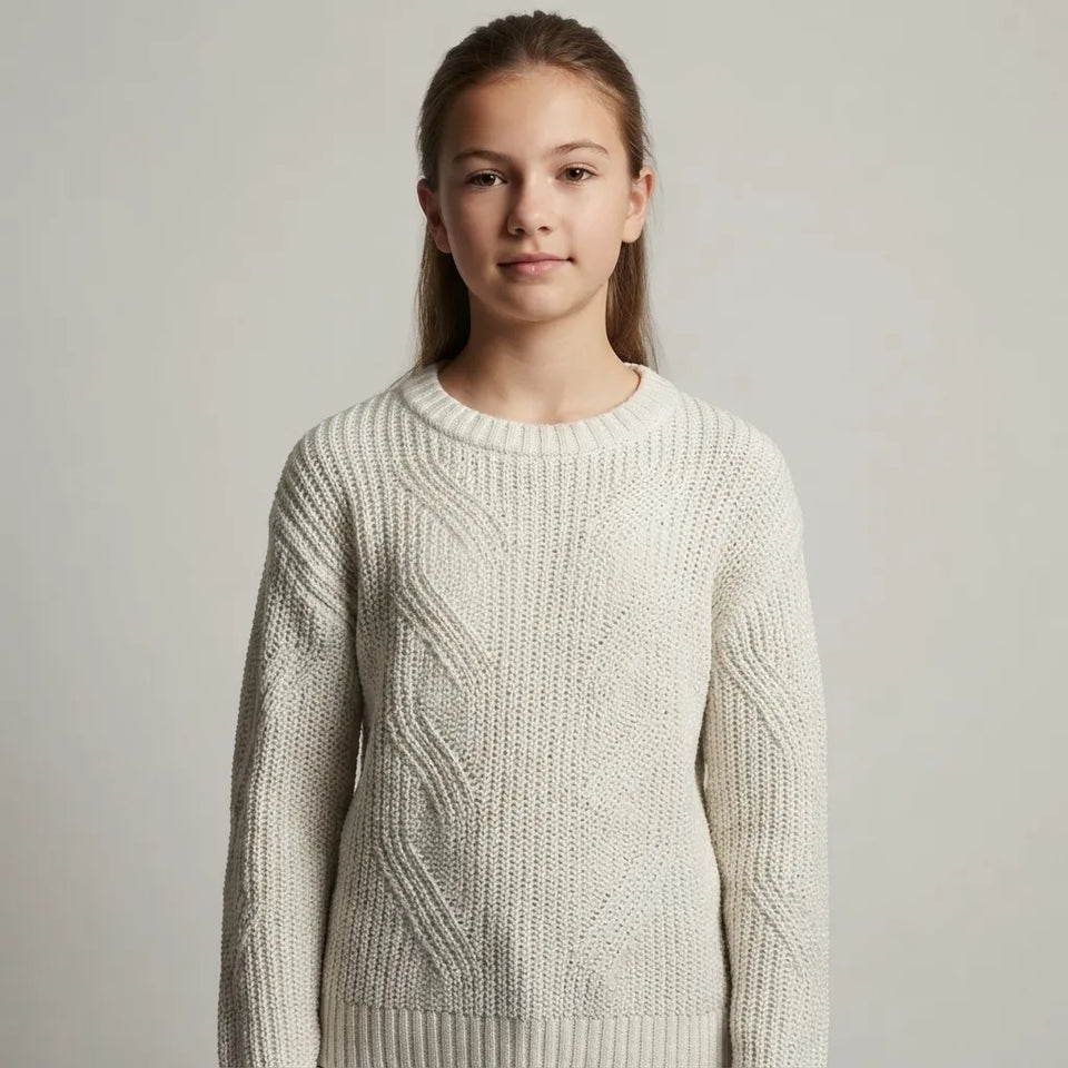A young girl with light brown hair tied back, wearing a cream-colored cable-knit crewneck sweater, photographed against a neutral light gray studio background.