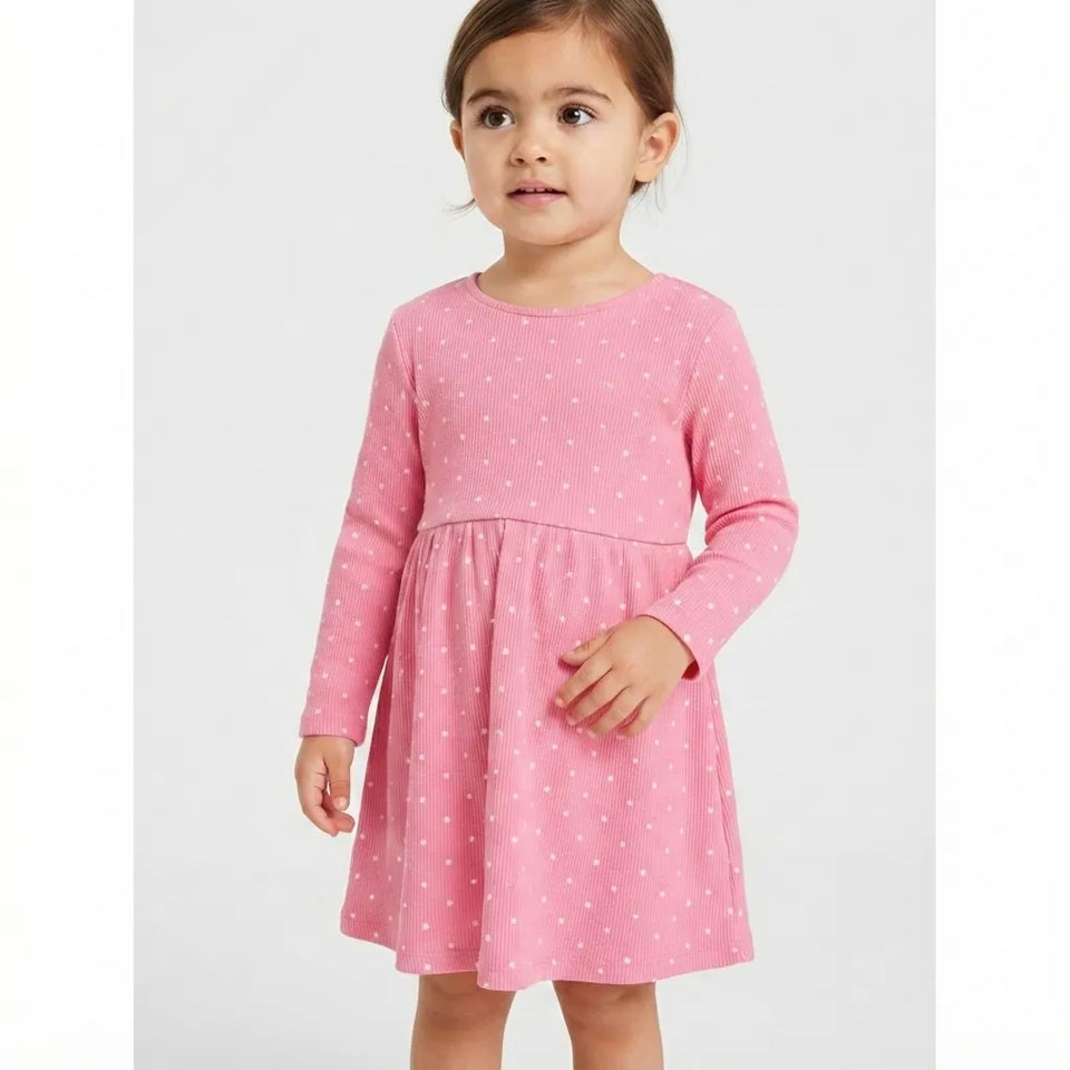 A young girl wearing a pink long-sleeve dress with a white polka dot pattern, standing against a light gray studio background.