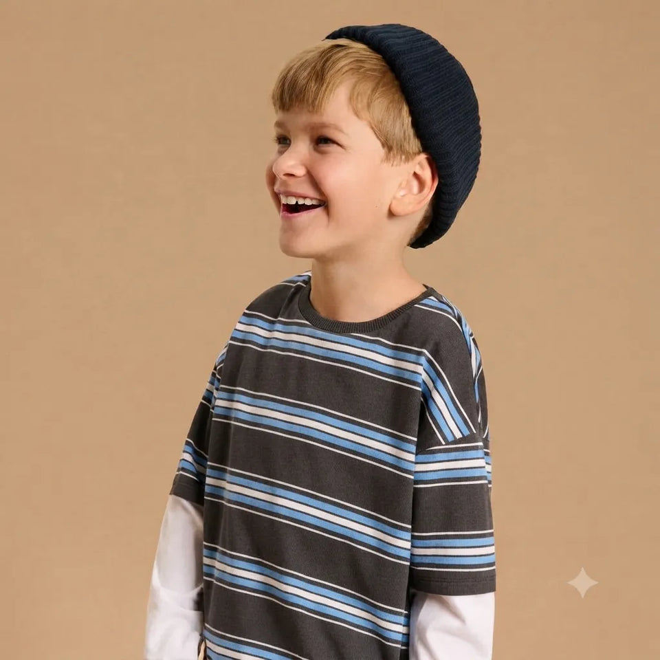 A young boy smiling and looking to the side, wearing a dark navy knit beanie and a dark gray striped t-shirt with blue and white stripes over a white long-sleeve shirt, set against a solid beige background.