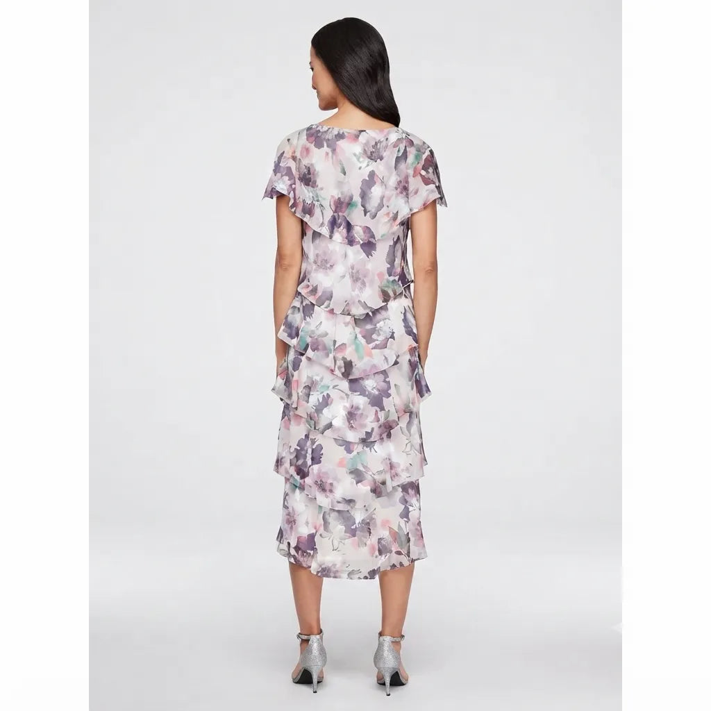 A woman viewed from the back wearing a short-sleeve midi dress with a ruffled neckline and tiered skirt, featuring a soft watercolor floral print in muted pink, purple, and gray tones. She is standing against a light gray studio background.