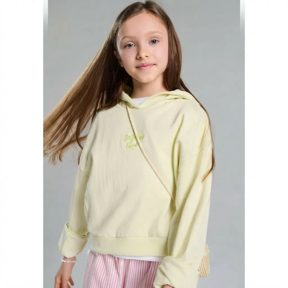 A young girl with long brown hair wearing a pale yellow-green hooded sweatshirt with a small embroidered logo on the chest, paired with pink and white striped pants, set against a plain gray studio background.