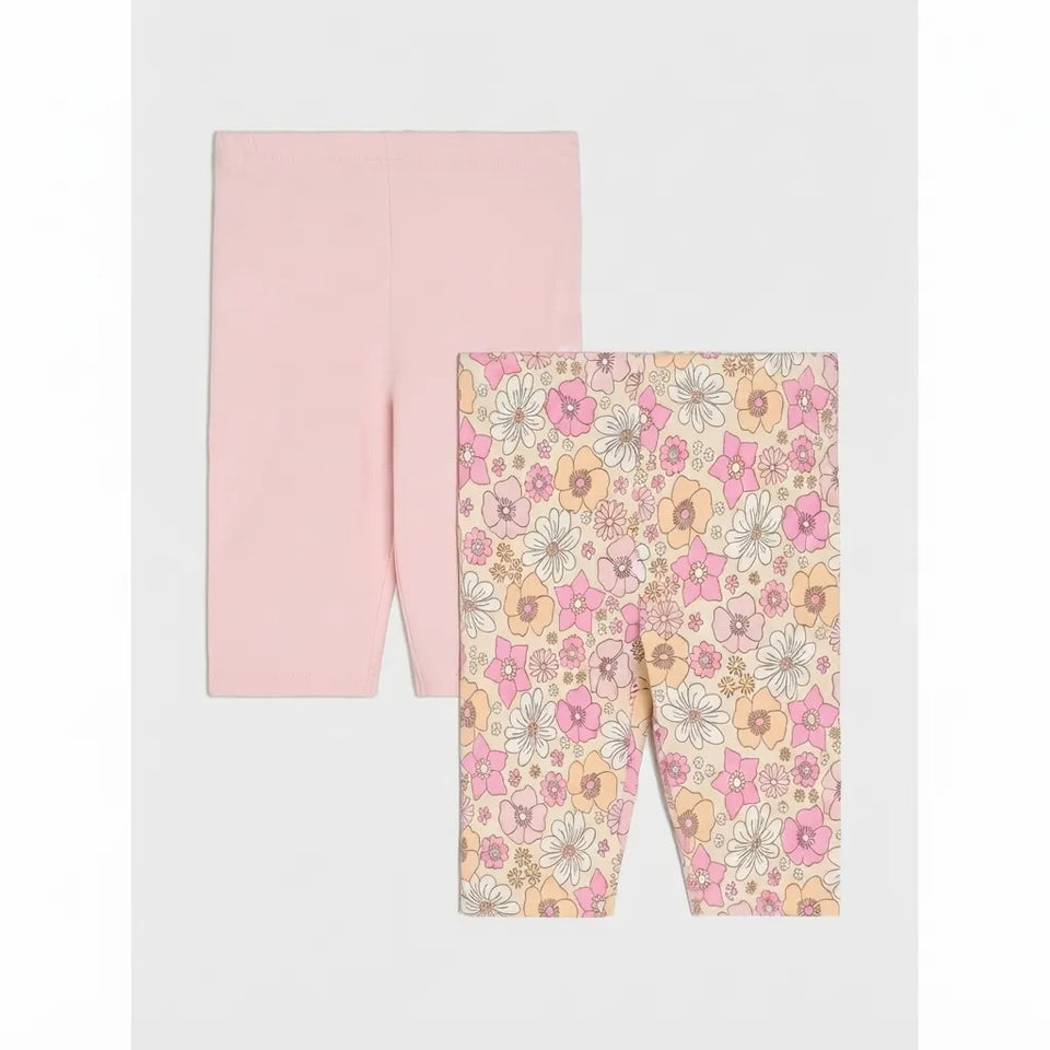 Two pairs of children's leggings laid flat on a light gray background. The back pair is solid light pink, and the front pair has a dense floral print with pink, peach, white, and gray flowers on a pale beige background.