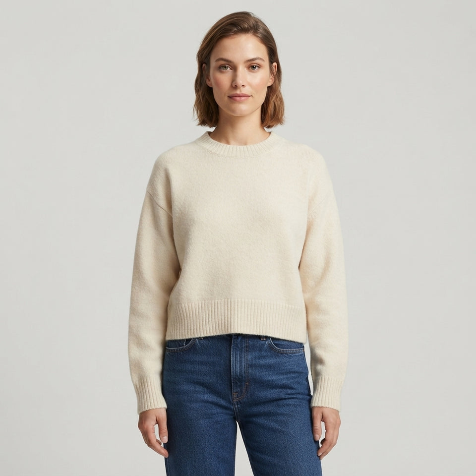 A woman wearing a cream-colored crewneck knit sweater and dark blue jeans, standing against a light gray studio background.