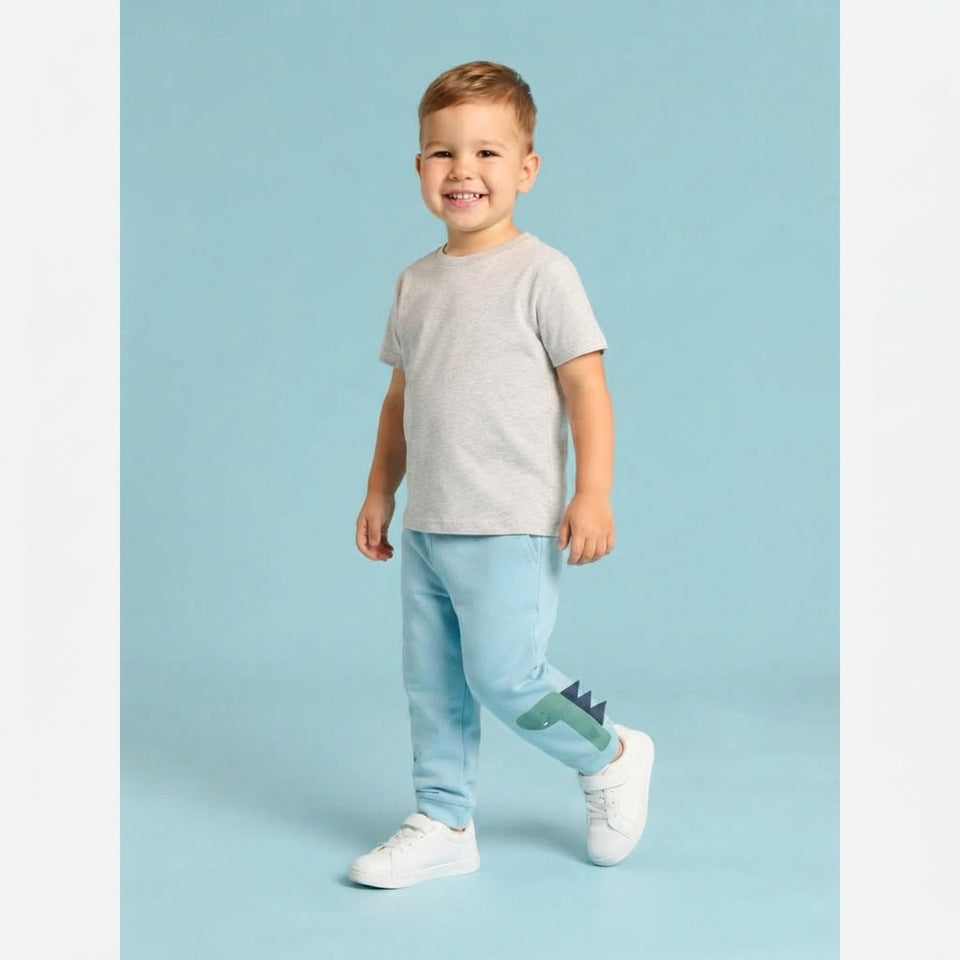 A toddler boy wearing a light gray short-sleeve t-shirt, light blue jeans with a green dinosaur graphic on the side, and white sneakers, standing against a solid light blue background.
