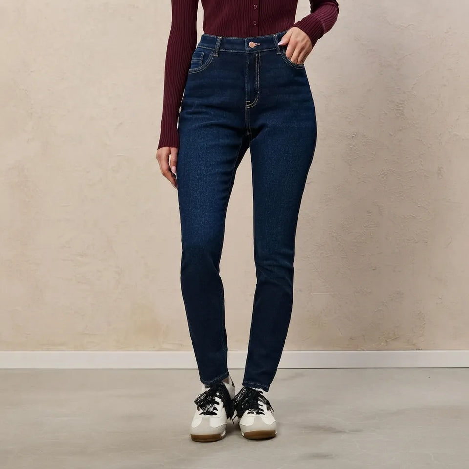 A model wearing dark blue high-waisted skinny jeans, a burgundy ribbed long-sleeve top, and white sneakers, standing against a textured beige wall.