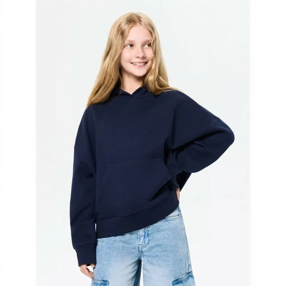 A young girl with blonde hair wearing a dark navy blue oversized pullover hoodie and light wash blue jeans, standing against a plain white studio background.