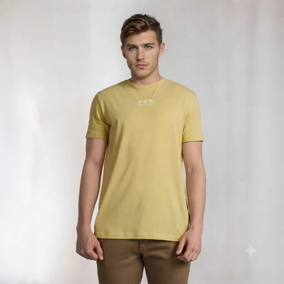 A man wearing a pale yellow crew-neck t-shirt with a small white logo on the chest, paired with brown pants, standing against a light gray studio background.